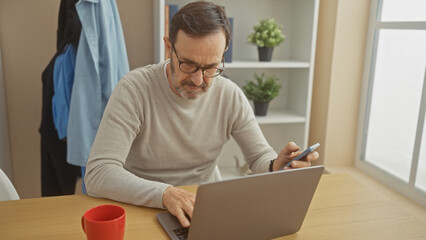 Bearded middle-aged man using a smartphone and laptop at a home desk by a window.