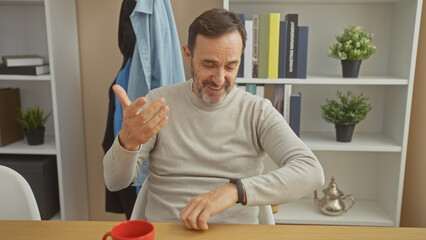 Middle-aged bearded man in sweater smiling while checking time on wristwatch at home, with...