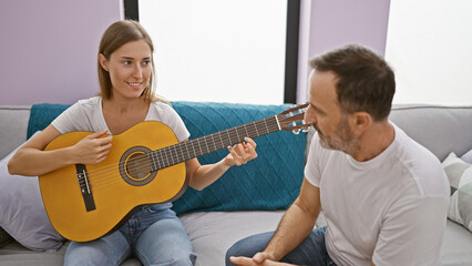 Father and daughter bonding while sitting on the sofa at home, playing classical guitar; their...