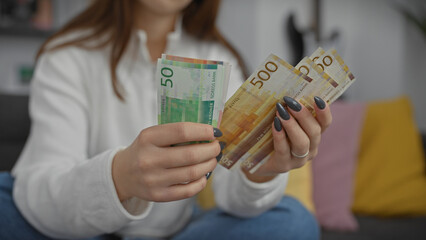 A young woman indoors holds norwegian kroner in a cozy home setting, suggesting wealth or budgeting.