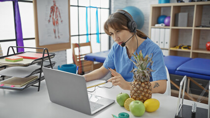 Focused caucasian woman in scrubs consulting on her laptop with a headset in a physiotherapy clinic...