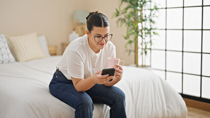 Young beautiful hispanic woman using smartphone sitting on the bed at bedroom