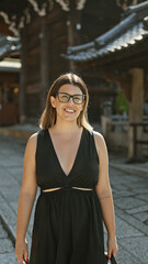 Confident hispanic woman, beautiful and joyful, cheerfully posing with glasses on, standing and smiling on kyoto's traditional streets, radiating natural and carefree latin expression.