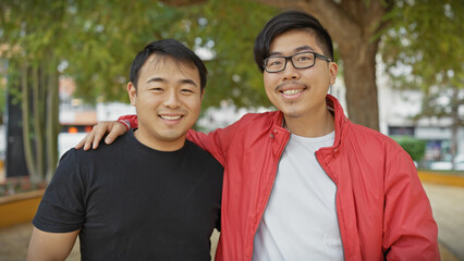 Two asian men smiling together in a park setting with green trees in the background, portraying...