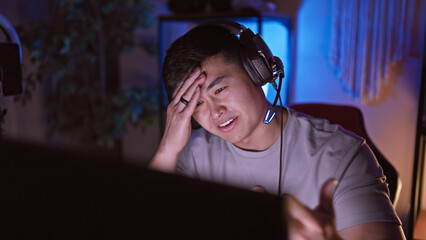 Frustrated asian man with headphones in a dark gaming room showing emotions of defeat or challenge.