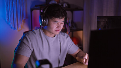 A young asian man focused on a screen in a dark gaming room wearing headphones at night.