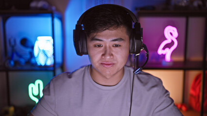 A young asian man wearing headphones indoors in a dark room illuminated by colorful gaming lights.
