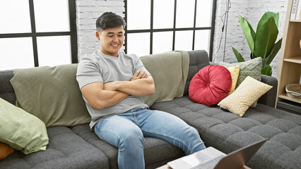 A young asian man relaxes on the sofa at home in a contemporary living room with bricks and plants.