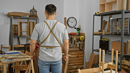 Back view of a young asian man standing thoughtfully in a well-organized carpentry workshop.