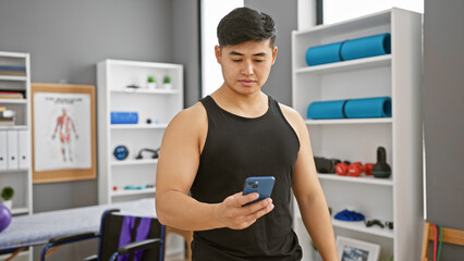 A young asian man in a sleeveless top using a smartphone in a physiotherapy clinic's well-equipped room.