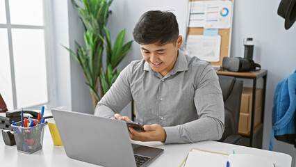 A young asian man smiles while using a smartphone at his modern office desk, portraying a professional, work environment.