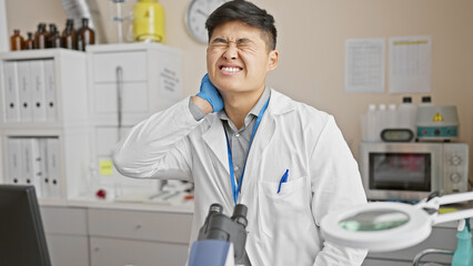A young asian man in a lab coat grimaces, holding his neck in pain, indicating discomfort inside a modern medical laboratory.