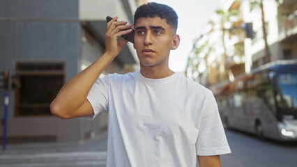 A young man listens to a voice message on his phone in the urban environment of a city street.