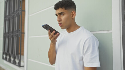A young adult man speaking into a smartphone on a city street, looking focused and engaged.