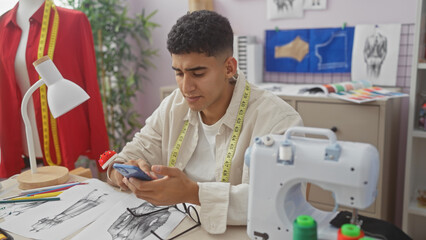 A young man using a smartphone in a tailor shop surrounded by sewing equipment and fashion design sketches.
