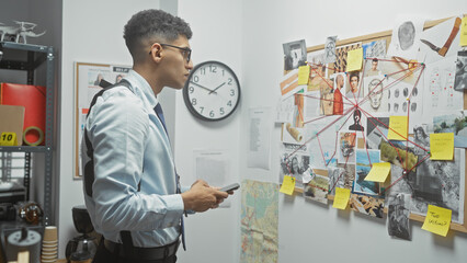 A young man analyzes a crime board with photos and notes in a detective's office indoors.