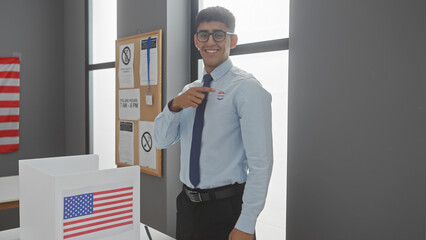 A smiling young man pointing at his 'i voted' sticker in an american polling station with a us flag.