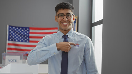 A smiling young man points to his 'i voted' sticker in a us polling station with an american flag in the background.