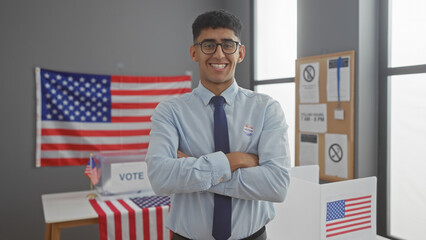 Young man with crossed arms smiling in an american voting center, adorned with flags and a voting booth.