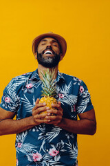 handsome mid adult african american man meditating with eyes closed and Hawaiian shirt posing holding pineapple to his chest on yellow background
