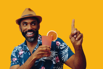 happy bearded mid adult african american man wearing Hawaiian shirt and hat smiling and orange juice cocktail pointing up and looking away at copy space on yellow background