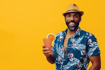 handsome bearded mid adult african american man wearing Hawaiian shirt and hat smiling with orange juice cocktail looking at camera on yellow background