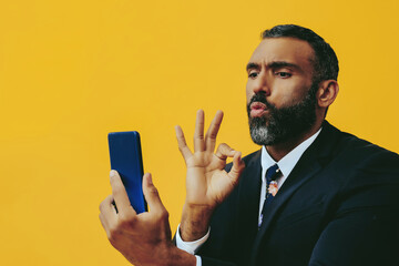 portrait of smiling positive handsome bearded man in suit and tie with smartphone video call ok sign hand on yellow background