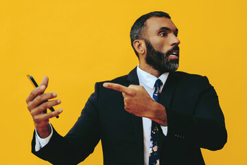 portrait of angry expressive excited bearded man in suit and tie with smartphone video call pointing finger at camera yellow background