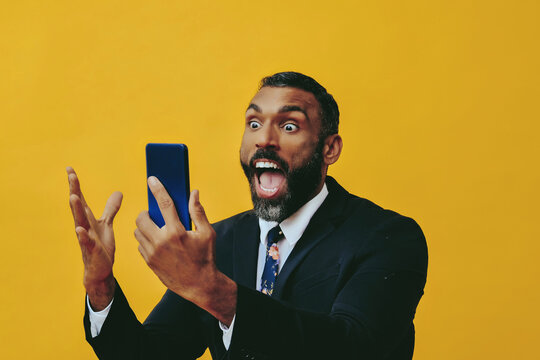 portrait of expressive angry excited bearded man in suit and tie with smartphone video call hand up yellow background