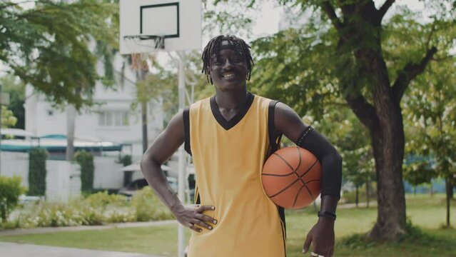 Portrait of cheerful black streetball player holding ball while posing for camera on outdoor playground