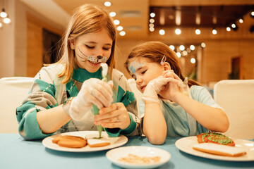 Two girls with aquagrim on their faces decorate Christmas cookies with a pastry bag in a cafe or restaurant. Sisters in casual clothes have fun and paint ginger sweetness with cream.