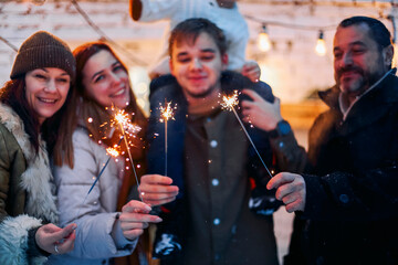 Portrait of big happy family with child standing in snow-covered house yard holding lit sparklers...