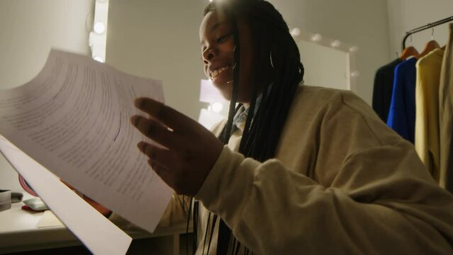 Low angle of humorous African American woman with braided hairstyle sitting in front of mirror with light bulbs in dressing room and laughing at jokes, preparing to give standup monologue on stage