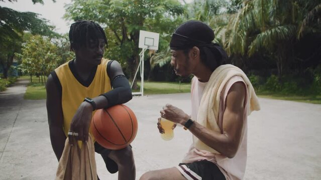 Medium long shot of black basketball player drinking refreshing beverage from bottle and chatting with team member on outdoor playground