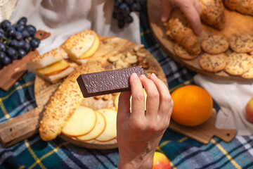 Close-up of hand holding chocolate against a background of laid out picnic food, selective focus. The concept of summer outdoor recreation on the weekend