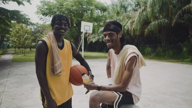 Medium long portrait of two black male streetballers in sportswear smiling at camera on outdoor playground