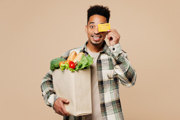 Young man wears grey shirt hold paper bag for takeaway mock up with food products cover eye with credit bank card isolated on plain pastel beige background. Delivery service from shop or restaurant.