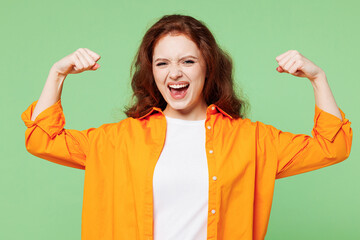 Young ginger woman she wears orange shirt white t-shirt casual clothes showing biceps muscles on hand demonstrating strength power isolated on plain pastel light green background. Lifestyle concept.