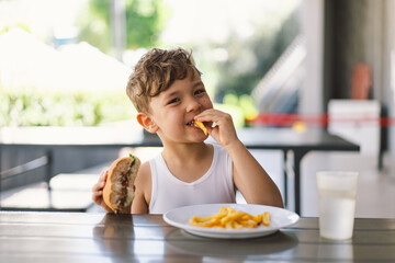 Little Boy Eating Sandwich and French Fries at Table - obrazy, fototapety, plakaty