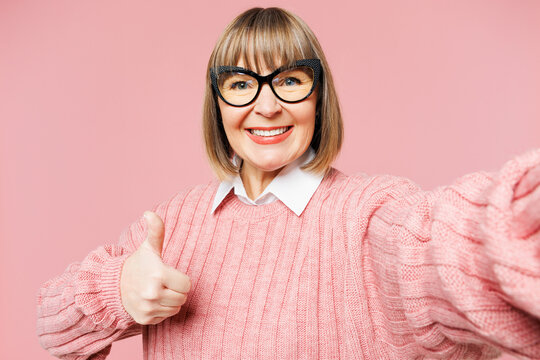 Close up elderly woman 50s year old wear sweater shirt casual clothes glasses do selfie shot pov on mobile cell phone show thumb up isolated on plain pink background studio portrait Lifestyle concept