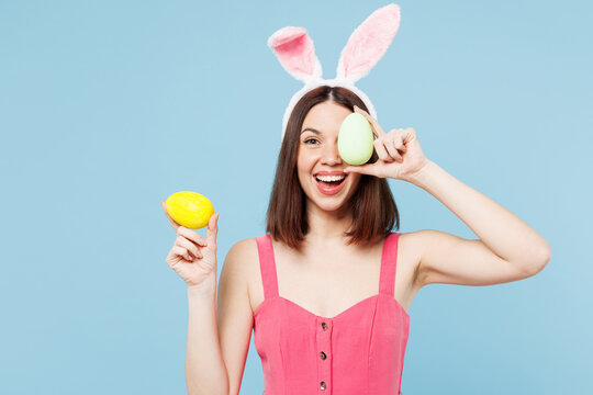 Young shocked cheerful woman wear pink casual clothes rabbit bunny ears hold in hand cover eye with painted eggs isolated on plain blue cyan background studio portrait. Lifestyle Happy Easter concept.