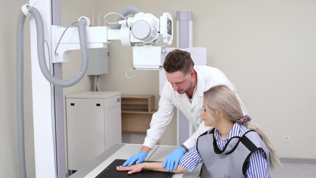 Man doctor in white medical uniform adjusts X Ray machine for scanning patient in clinic hospital. Radiologist and patient in a x-ray room. X-ray of human hand. Classic ceiling-mounted x-ray system. 