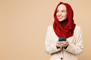 Young Arabian Asian Muslim woman wear red abaya hijab suit clothes hold use mobile cell phone look aside on area isolated on plain beige background studio. UAE middle eastern Islam religious concept.