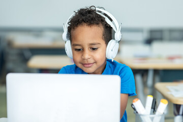 Primary school boy student with headphones using laptop, typing on keyboard, sitting in classroom interior