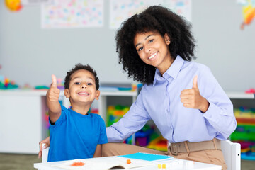 Tutoring and mentorship concept. Black female teacher and happy schoolboy gesturing thumbs up and smiling sitting at desk in class