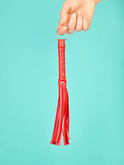 Red whip for adult role play games in woman's hand over turquoise blue background - 785414320