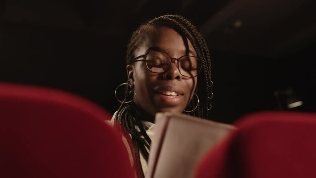 Chest up of young talented Black female actress reading play script aloud sitting on red seat in dark theater during rehearsal or auditions