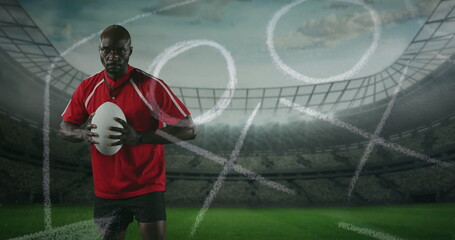 Image of an African American male rugby player standing, playing with a ball and looking to camera w