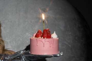 girl hand holds glamorous pink strawberry birthday cake specially for woman with burning candle on it. Make a wish, blow a candle. Starlight effect on glowing flame, blazing star. Celebration day