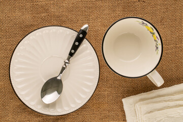 One white porcelain cup with saucer, spoon and napkin on jute cloth, macro, top view.